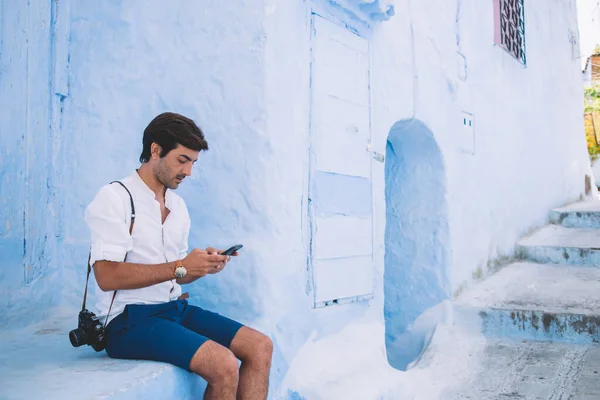 Side view of millennial hipster guy in casual wear using smartphone gadget for online phoning while sitting near ancient chaouen buildings, amateur photographer with retro camera sending sms