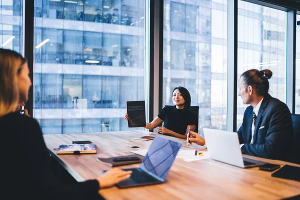 Elegant japanese woman in formal wear showing graphic with productive strategy for company working, group of business people collaborating on accounting in modern office sitting at desk with laptops