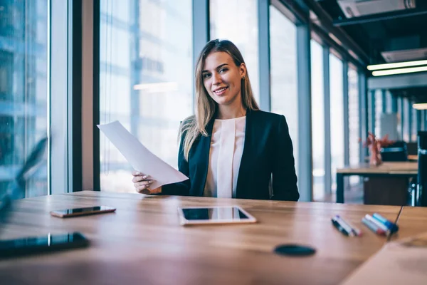 Portrait of successful caucasian female entrepreneur sitting at desktop with paper documents and looking at camera while working on strategy for good organisation of business event with partners