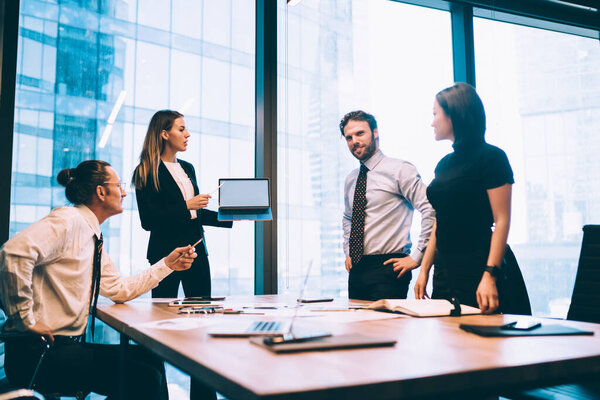 Intelligent female employee in elegant suit holding modern laptop computer with mock up area for advertising text for financial website while having brainstorming process with professional colleagues