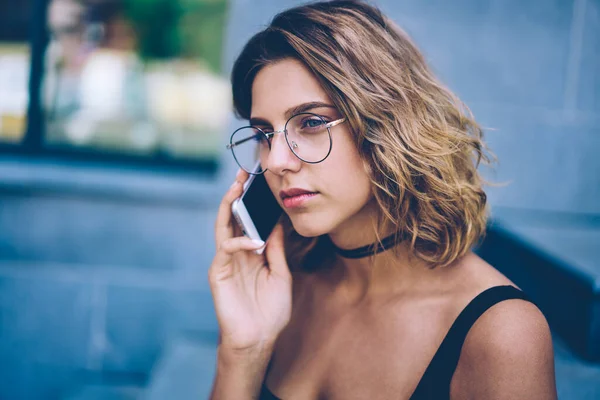 Serious hipster girl calling to operator for consultancy about tarification for communicate in roaming, attractive female teenager in sunglasses making mobile conversation via cellular gadget