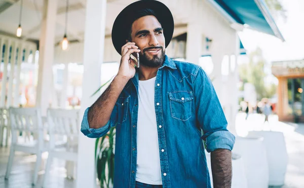 Positive man in fashionable black hat smiling during smartphone cell call with roaming internet connection, successful male millennial in trendy apparel communicating via digital mobile phone