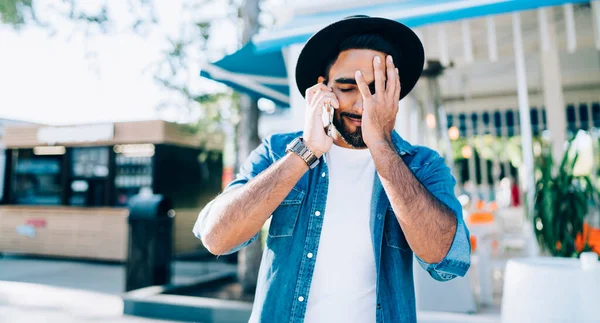 Frustrated hipster guy in stylish apparel feeling sad from making mistakes with using money in roaming from account balance phoning to operator for online consultancy, concept of memory reminder