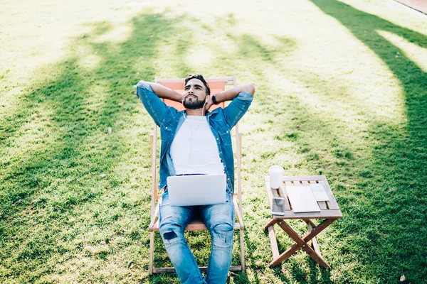 Top view of handsome Turkish man dressed in casual denim apparel enjoying beauty of life and summer weather, millennial hipster guy taking rest outdoors listening positive audio book using laptop