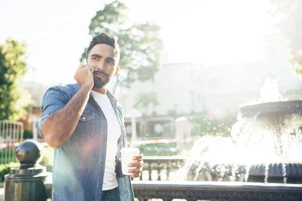 Turkish hipster guy with caffeine beverage in hand standing on city street during sunny summer day and using roaming internet connection for making cellphone conversation with operator for consultancy