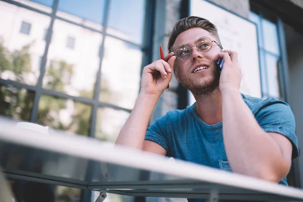 Contemplative young man in spectacles for vision correction pondering information during consultancy conversation with operator, pensive focused male thinking hard while speaking on cellphone