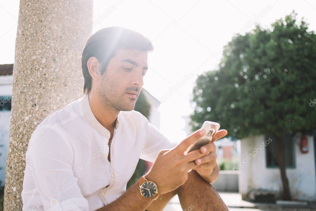 Young Hindu hipster guy sitting at urban setting and searching information on website connected to 4g wireless on cellphone gadget for networking, concept of digital technology and communication
