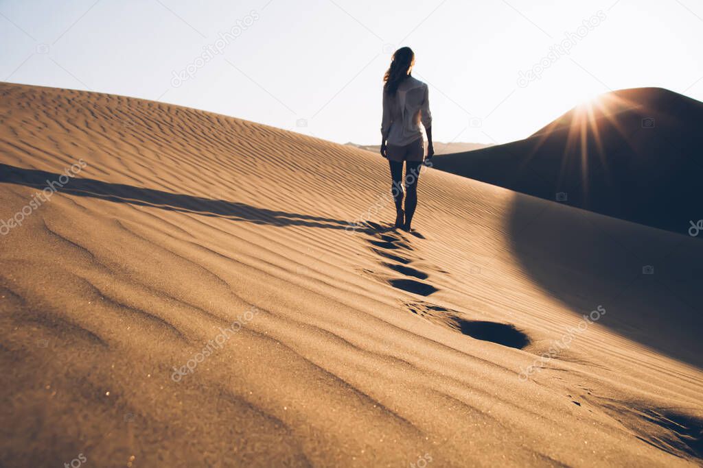 Back view of female wanderer in casual wear exploring wild Sahara desert during summer evening in nature environment, woman tourist walking in sands wilderness discovering hot Egypt climate
