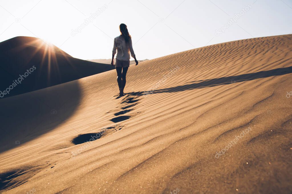 Back view of female wanderer in casual wear exploring wild Sahara desert during summer evening in nature environment, woman tourist walking in sands wilderness discovering breathtaking Safari