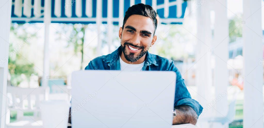 Portrait of cheerful Turkish software developer sitting at desktop with laptop device and smiling at camera while waiting for technology updating, happy man in casual wear working remotely