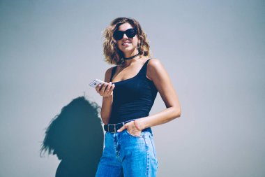 Akıllı telefon zımbırtısıyla kameraya bakan mutlu milenyum hipster kızının yarım boy portresi. Güneş gözlüklü pozitif bir kadın boş vakitlerinde elinde cep telefonu tutuyor.