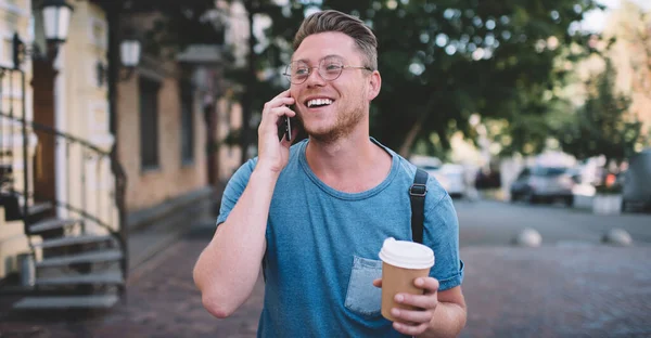 Excited Caucasian hipster guy with takeaway coffee in hand satisfied with friendly smartphone conversation during time for travelling, happy cheerful man in optical spectacles smiling while phoning