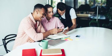 Başarılı erkek ve bayan meslektaşlar, interneti cep telefonuyla taramak için işe ara vererek işe ara veriyor, neşeli çeşitli öğrenciler öğrenme ve gülümseme teçhizatlarıyla masaüstünde oturuyor