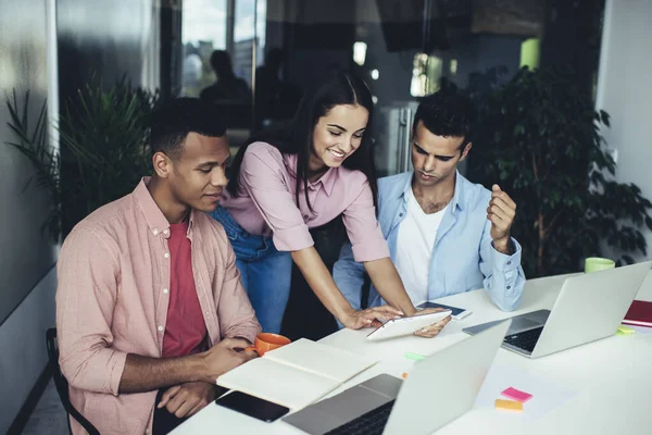 Cheerful female employee smiling and showing funny video to diverse male colleagues for watching connected to 4g wireless in office, group of happy entrepreneurs reading news in social networks