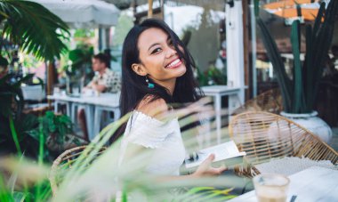 Beyaz omuzlu, kitap okuyan, terasta bir fincan kahveyle oturmuş gülümseyen Asyalı güzel bir kadının yan görüntüsü.