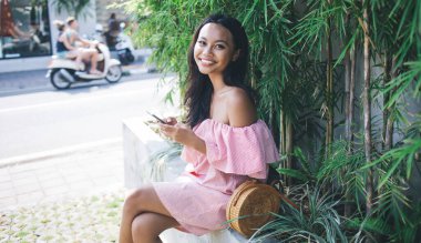 Yeşil tropikal yapraklarla kaplı duvarda otururken pembe omuzlu güzel Asyalı kadının akıllı telefonuyla göz gezdirişinin yan görüntüsü