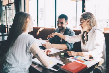 Bir grup modern genç kadın ve erkek masada kağıt ve çalışmayla birlikte otururken aynı zamanda takım halinde yeni bir proje üzerinde çalışıyorlar.