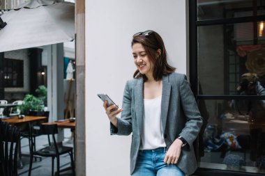 Güzel, güzel, mutlu, resmi giyinmiş, akıllı telefon kullanan, şehrin sokaklarında duvara yaslanmış, gülen bir kadın.