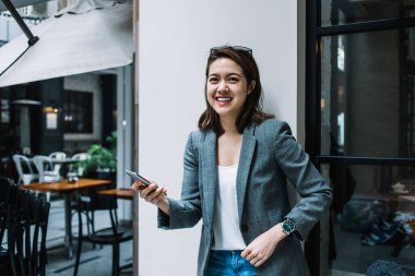 Genç, çekici, güzel, neşeli, resmi giyinmiş, akıllı telefon kullanan, duvara yaslanmış, caddede kameraya bakan bir kadın.