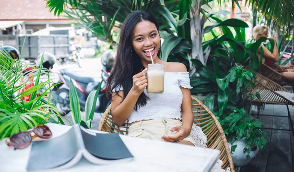 Cheerful pretty young Asian woman in off-shoulder blouse sipping cold coffee from mason jar through straw while sitting at table in open terrace