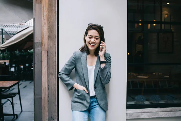 Young beautiful cheerful ethnic woman in formal clothes speaking on smartphone with closed eyes with joy laughing on city street