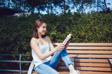 Kotlu rahat bir etnik kadın yazın güneşli bir parkta ahşap bankta oturup ilginç bir kitap okuyor. 