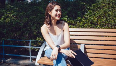 Sırıtkan Asyalı genç bayan kitapla, kolları çapraz ve kısa kahverengi saçlarla tahta bankta otururken diğer tarafa bakarken çitlere doğru bakıyor.