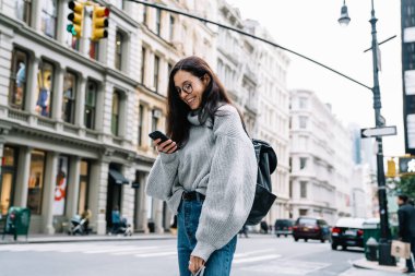 Gri kazaklı, mavi kotlu ve gözlüklü genç bir bayan kaldırımda duruyor. New York 'ta alışveriş torbaları taşıyor ve akıllı telefondan mesajlaşıyor.