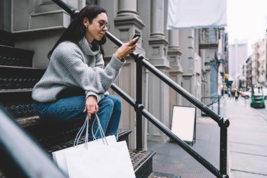 New York 'un caddesinde akıllı telefonlar üzerinde sörf yaparken elinde boş beyaz alışveriş torbalarıyla merdivenlerde oturan günlük kıyafetli ve gözlüklü genç bir kadının yan görüntüsü. 