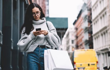 Daha şık giyinmiş, gözlüklü, elinde alışveriş torbalarıyla New York sokaklarında yürürken akıllı telefondan mesajlaşan güzel bir yetişkin kız.
