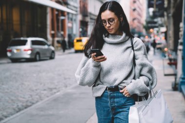 Gözlüklü, kot pantolon ve süveter giyen çekici çağdaş kadın. Şehir caddesinde faiziyle akıllı telefon kullanıyor.
