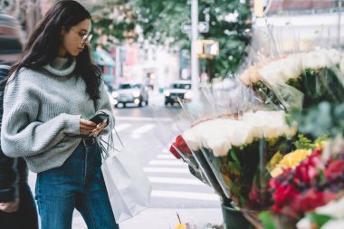Gri kazaklı ve gözlüklü bir kadın elinde alışveriş torbalarıyla kaldırımda duruyor ve New York 'ta sokakta telefonla dikiliyor.