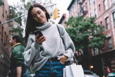 Kazaklı, gözlüklü, elinde alışveriş çantası olan ve New York 'ta sokakta yürürken cep telefonuyla gezen mutlu modern bir kadın.