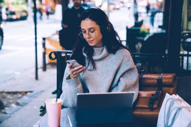 Gözlüklü, kulaklıklı, akıllı telefonlu, modern tabletli modern New York 'un kafe terasında oturan genç ve güzel bir bayan. 