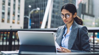 Kendine güvenen, gri ceketli ve gözlüklü iş kadını New York şehrinin terasındaki tableti incelerken şehir merkezindeki masada oturmuş belgeleri kontrol ediyor.