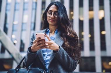 Deri çantalı, gözlüklü, gözlüklü, şehrin arka planında telefonla konuşan etnik bir kadından.