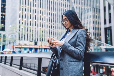 Deri çanta ve cep telefonuyla şehrin arka planında duran mesajlar taşıyan gözlüklü ve modaya uygun ceketli modern etnik bayan.
