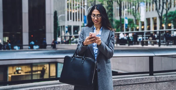 眼鏡やコートの現代的な流行の民族の女性は 革のブリーフケースを保持し ニューヨーク市を背景に携帯電話でテキストメッセージ — ストック写真