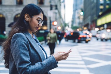 Odaklanmış, yuvarlak gözlüklü, gri montlu, New York 'un kalabalık caddesinde, bulanık arka planda yaya geçidinde akıllı telefondan konuşan esmer bir kadının yan görüntüsü.