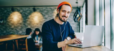 Mutlu gülümseyen hipster adamın portresi kafeteryada öğrenim görürken favori müzik listesi için zaman harcıyor, kulaklıklı pozitif bir genç evde dizüstü bilgisayarla masada oturuyor.