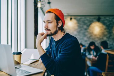 Elektronik kulaklıkla ilginç ses kitaplarını dinlerken şık giyinmiş düşünceli hippi adam dizüstü bilgisayar için 4G internet hakkında kafa yoruyor.