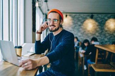 Optik gözlüklü olumlu erkek meloman, dost canlısı bir sesli mesajla kendini iyi hissediyor, kafeteryada dizüstü bilgisayarla oturan ve kulaklıkla müzik listesi dinleyen mutlu hipster adam.
