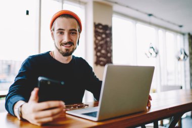 Akıllı telefon cihazını elinde tutan olumlu hipster adamın portresi öğrenme ve kameraya gülümseme için dizüstü bilgisayarla masada otururken, mutlu erkek serbest çalışan ve birlikte çalışma konusunda kendini iyi hisseden