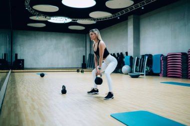Son moda spor kıyafetli genç bir kadının Kettlebell 'le spor yapıp aynaya bakarken spor salonunda çömelmesi.