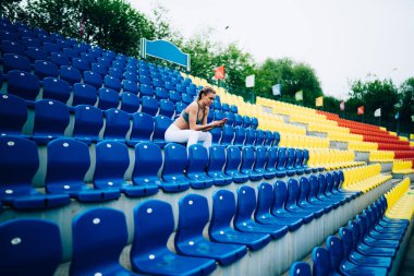 Spor giyimli yetişkin bir kadının renkli stadyumda oturduğu ve yeşil ağaçlara karşı akıllı telefonlara baktığı yan manzara. 