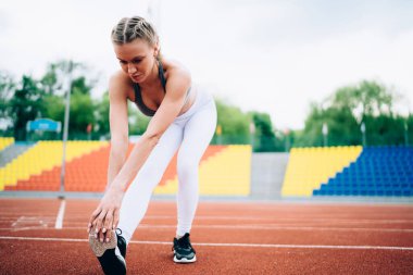 Spor giyimli, sarışın, aktif genç bir kadın yazın stadyum pistinde ayak parmaklarına yaslanırken bacak kaslarını esnetiyor.
