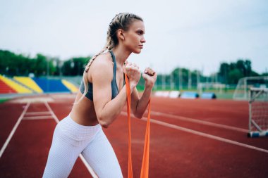 Açık hava stadyumunda kol kasları açmak için kauçuk jimnastik bandı geren uzun sarı saçlı, konsantre spor kadın.