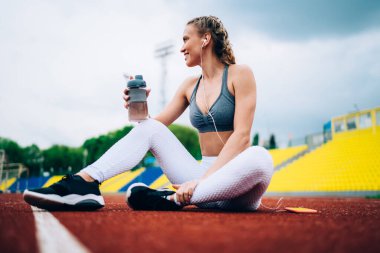 Akıllı telefondan müzik dinleyen, elinde su şişesi tutan ve boş stadyumun koşu yolunda oturan ve gözlerini kaçıran gülümseyen bayan sporcunun zemin katındaki görüntüsü.