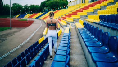 Modern spor giysileri içinde ince bir kadın ve örgülü saçlarıyla stadyumdaki renkli tribünlerin arasında yürüyor.