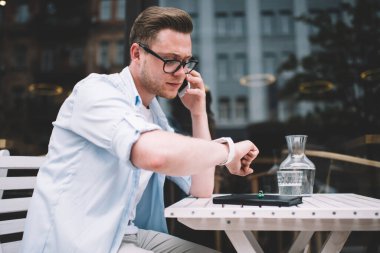 Bin yıllık beyaz gözlüklü genç bir adamın cep telefonuyla dışarıdaki masada oturan kol saatine bakarken görüntüsü.